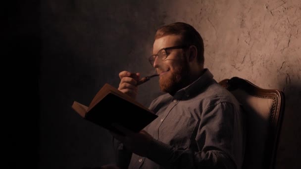El hombre lee un libro en un cuarto oscuro y fuma pipa. 4K UHD — Vídeo de stock
