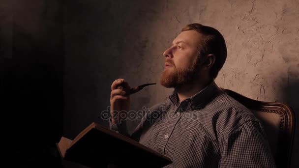 O homem lê um livro num quarto escuro e um cachimbo de fumo. 4K UHD — Vídeo de Stock