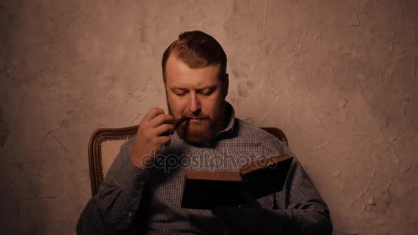 O homem lê um livro num quarto escuro e um cachimbo de fumo. 4K UHD — Vídeo de Stock