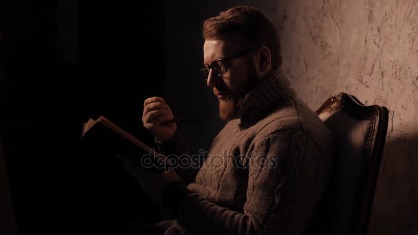 O homem lê um livro num quarto escuro e um cachimbo de fumo. 4K UHD — Vídeo de Stock