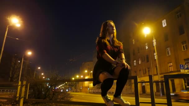 Chica adolescente lindo moderno sentado por la noche en una ciudad y disfrutar de los días cálidos de principios de primavera. Luz ambiental, carretera y coches sobre un fondo . — Vídeo de stock