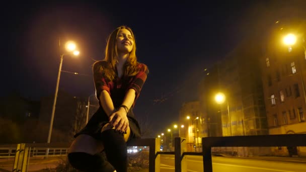 Moderne schattige tiener meisje zit bij nacht in een stad en geniet van de warme dagen van de vroege lente. Omgevingslicht, weg en auto's op een achtergrond. — Stockvideo