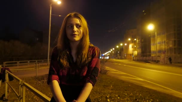 Moderna ragazza adolescente carina seduta di notte in una città e godersi le calde giornate di inizio primavera. Luce ambientale, strada e auto su uno sfondo . — Video Stock