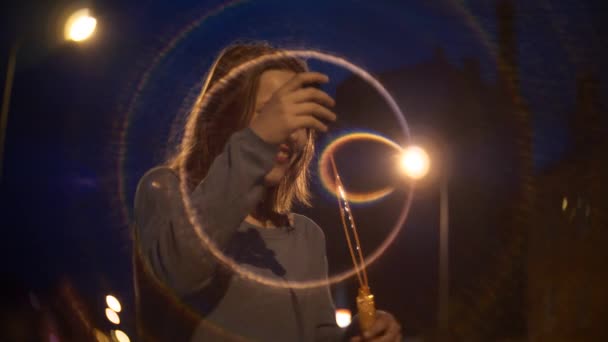 Портрет милої усміхненої дівчини-підлітка на нічній міській вулиці, що дме мильні бульбашки — стокове відео