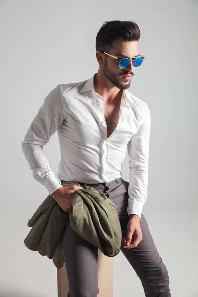 Zittende jonge mode man met zonnebril holding zijn jasje — Stockfoto