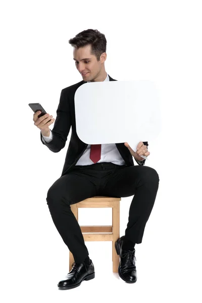 Businessman looking at his phone and holding a speech bubble — Stock Photo, Image