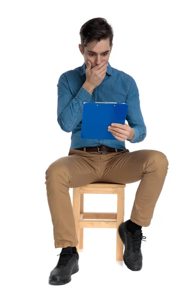 Shocked young man covering mouth and holding clipboard — Stockfoto