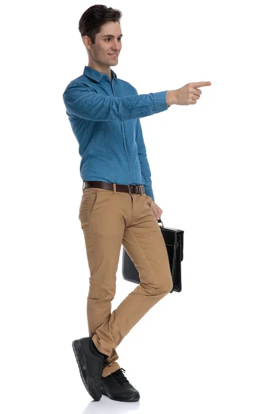 Mutlu genç adam bavulu tutuyor ve parmağıyla işaret ediyor — Stok fotoğraf