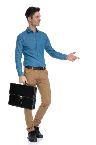 Joven elegante hombre sosteniendo la maleta y presentando a un lado — Foto de Stock