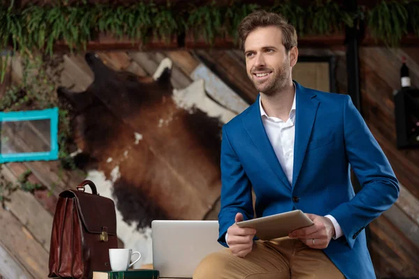 Hoopvolle zakenman die zijn tablet vasthoudt, wegkijkt en glimlacht — Stockfoto