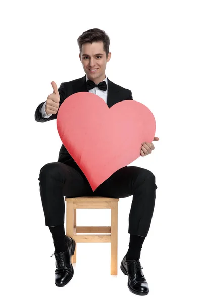 Happy young guy holding big red heart and making thumbs up sign — Stock Photo, Image