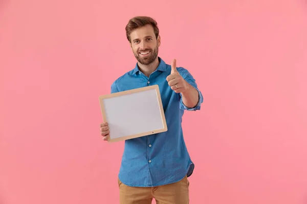 Šťastný mladý muž drží prázdnou desku a dělat palce nahoru znamení — Stock fotografie