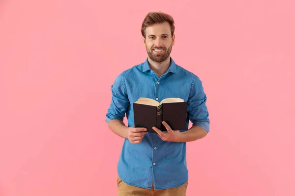 Feliz chico joven en camisa de mezclilla sonriendo y sosteniendo libro —  Fotos de Stock