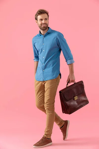 Улыбающийся молодой парень в джинсовой рубашке ходит и держит чемодан — стоковое фото