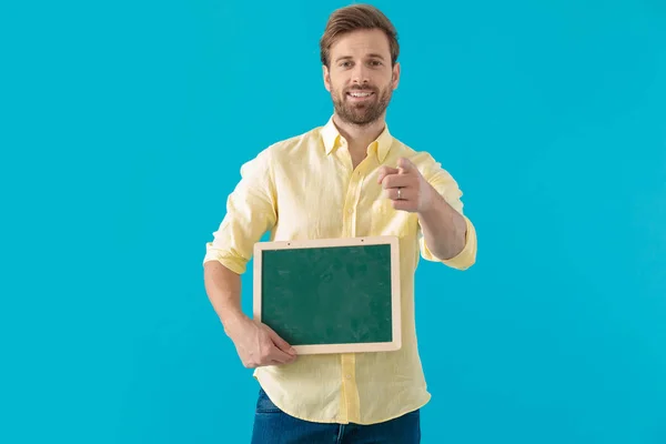 Positive casual man pointing forward and holding a blackboard — ストック写真
