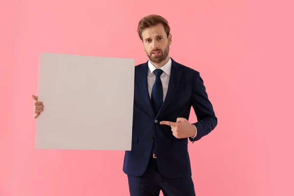 Unsure businessman holding a blank billboard and pointing to it — Stock Photo, Image