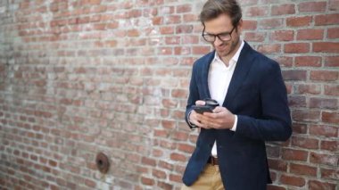 Mutlu genç işadamı kahvesini içiyor ve akıllı telefonuyla tuğla duvarın yanında geziniyor.