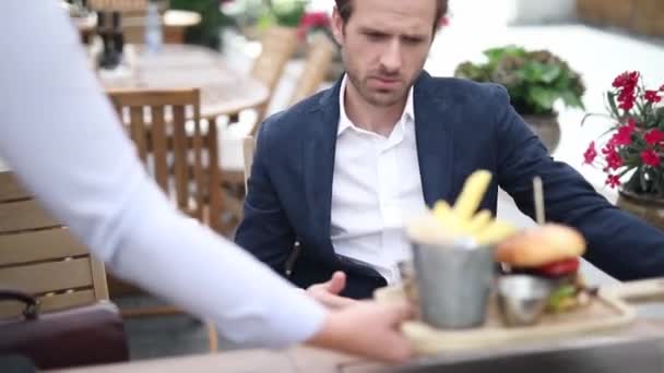 Νεαρός άνδρας που σερβίρει μεσημεριανό γεύμα σε ένα εστιατόριο μπιστρό - ο σερβιτόρος να του φέρει ένα φρέσκο μενού χάμπουργκερ με πατάτες — Αρχείο Βίντεο