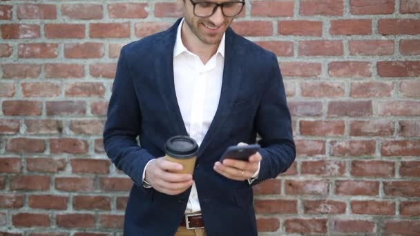 Jovem empresário feliz está bebendo seu café e lendo notícias em seu telefone inteligente perto da parede de tijolo — Vídeo de Stock