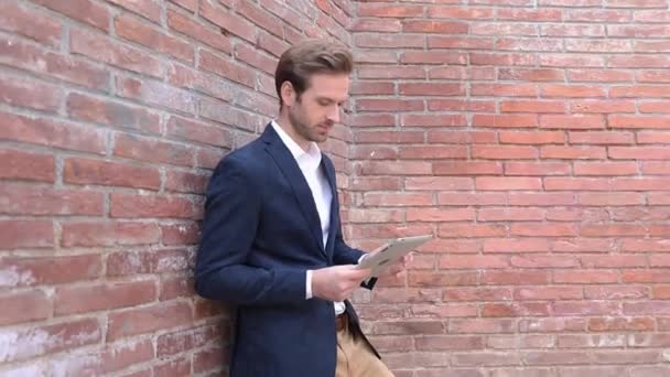 Felice uomo casuale intelligente appoggiato al muro di mattoni legge buone notizie su tablet e fa il pollice ok segno mano in su — Video Stock