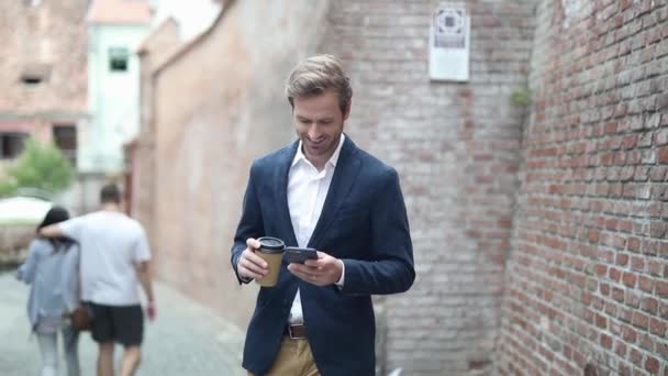 가볍게 걷고 커피를 마시고 있는 한 젊은 남자가 핸드폰으로 흥미 로운 뉴스를 읽고 있습니다. — 비디오