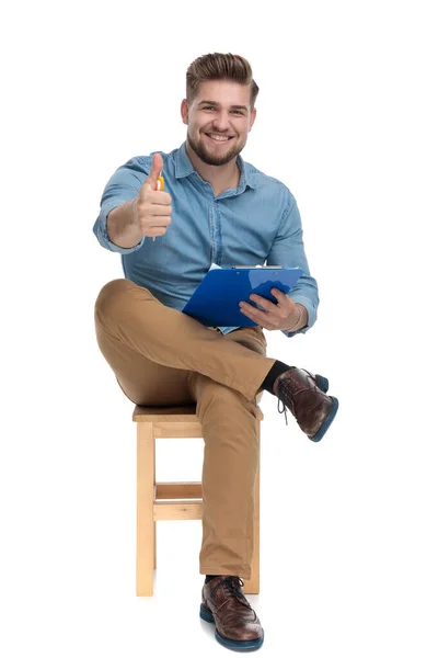 Happy young man making thumbs up sign and holding clipboard — Stockfoto