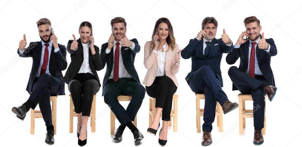 Team of 6 businessmen talking on phone and giving thumbs up while sitting on chairs on white studio background