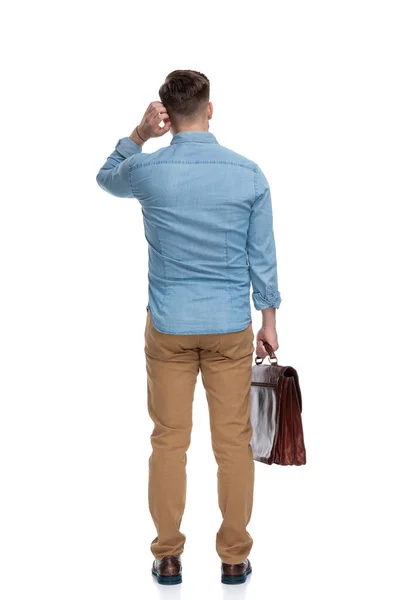 一个心事重重的漫不经心的男人抓着耳朵拿着公文包 站在白色的工作室背景下的后视镜 — 图库照片