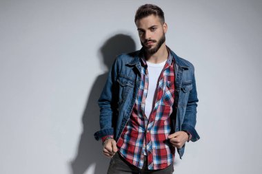 Gri stüdyo arka planında dururken ceketini ayarlayan yakışıklı bir manken.