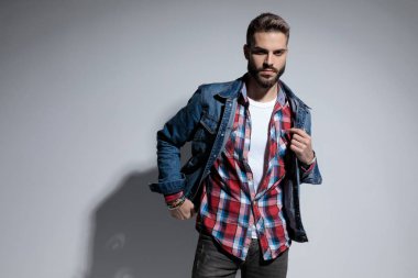 Gri stüdyo arka planında dururken ciddi bir genç model kaşlarını çatıyor ve ceketini tamir ediyor.