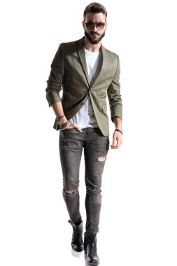 Yeşil ceket ve güneş gözlüğü giyerken elini cebinde tutan kendine güvenen bir model, beyaz stüdyo arka planında yürüyor.