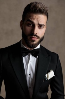 Yakışıklı damat smokin giyerken ve duvar kağıdı stüdyo arka planında bir taburede otururken ciddi bir şekilde ileriye bakıyor.