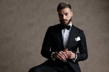 Düşünceli damat smokin giyerken ve duvar kağıdı stüdyo arka planında bir taburede otururken ileriye bakıyor ve merak ediyor.