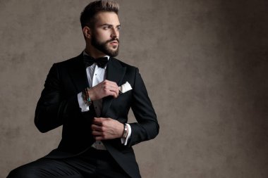 Düşünceli damat, smokin giyerken ve duvar kâğıdı stüdyo arka planında bir taburede otururken umutla yüzünü çeviriyor.