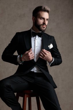 Kendine güveni olan damat smokin giyerken ve duvar kağıdı stüdyo arka planında bir taburede otururken ceketini ayarlıyor ve başka tarafa bakıyor.