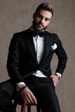 Smokin giyerken ve duvar kağıdı stüdyo arka planında bir taburede otururken elini cebine atmış meraklı damat.