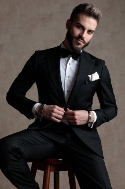 Kendine güveni tam bir damat smokin giyerken ceketinin düğmelerini açıyor ve duvar kağıdı stüdyosunun arka planında bir taburede oturuyor.