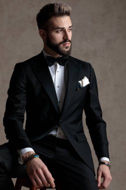 Meraklı damat smokin giyerken ve duvar kâğıdı stüdyo arka planında bir taburede otururken başka tarafa bakıyor.