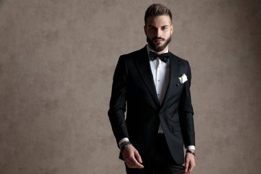 Genç damat smokin giyerken ve duvar kâğıdı stüdyo arka planında dururken ciddi bir şekilde ileriye bakıyor.