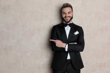 Neşeli damat, smokin giyerken ve arka planda duvar kağıdı üzerinde dururken kollarını kavuşturup gülüyor ve işaret ediyor.