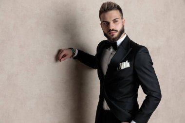 Smokin giyerken ve duvar kâğıdı stüdyo arka planında dururken elini cebine atmış sert bir damat.