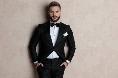 Sıkıntılı damat, smokin giyerken ve arka planda duvar kâğıdı üzerinde dururken iki elini cebine sokup ileriye bakıyordu.