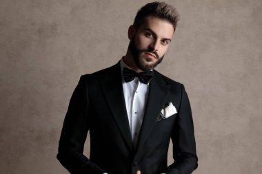 Yakışıklı damat smokin giyerken ve arka planda duvar kâğıdı üzerinde dururken ileriye bakıyor ve merak ediyor.
