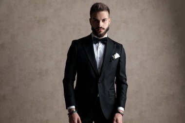 Gizemli damat smokin giyerken ve arka planda duvar kâğıdı üzerinde poz verirken ileriye bakıyor.
