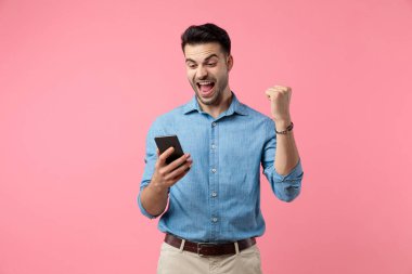 Sıradan bir genç adam bağırıyor ve telefona bakıyor, yumruğunu havaya kaldırıyor ve zaferi kutluyor, pembe arka planda duruyor.