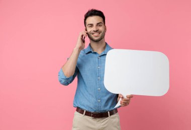 Genç adam elinde konuşma balonu tutuyor ve telefonda konuşuyor, pembe arka planda gülümsüyor.