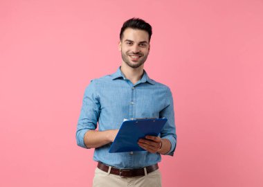 Gülümseyen, elinde not panosu olan, pembe arka planda duran mutlu, genç bir adam.