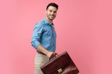 Gülümseyen, bavulu tutan, pembe arka planda yürüyen mutlu genç adam.