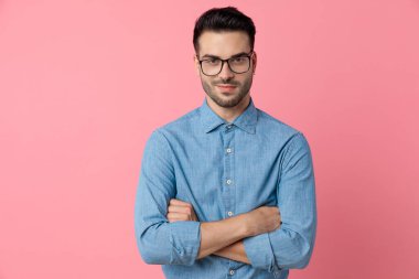 Kot gömlekli gülümseyen genç adam pembe arka planda kollarını kavuşturuyor.