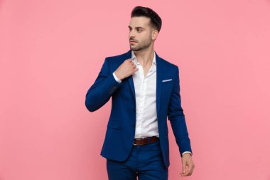 Yakasını düzelten ve mavi takım elbise giyip pembe stüdyonun arka planına basarak gözlerini kaçıran çekici bir iş adamı.